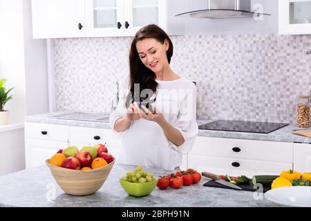 Smiling Young Woman Using Cellphone Near Fresh Fruits And Vegetables On Kitchen Counter Stock Photo