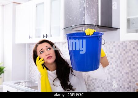 Worried Young Woman Calling Plumber On Cellphone And Collecting Water Leaking From Ceiling In Bucket Stock Photo
