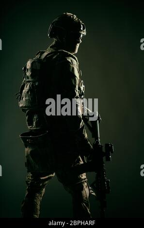 Low key studio shoot of army special forces soldier, commando fighter in mask, ballistic glasses, tactical helmet and battle uniform, holding short ba Stock Photo