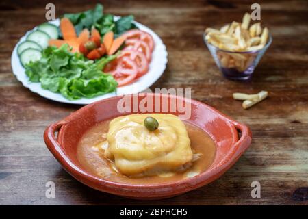 Portuguese francesine in a clay plate with salad and fried potatoes on a wooden background. Stock Photo
