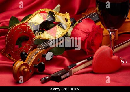 Violin (fiddle), theater mask, red heart, glass of wine and red rose lying on the perfect red satin fabric. String instrument. Valentine's day. Stock Photo
