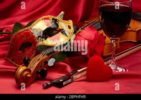 Violin (fiddle), theater mask, red heart, glass of wine and red rose lying on the perfect red satin fabric. String instrument. Valentine's day. Stock Photo