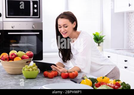 Happy Young Woman Using Digital Tablet Near Fresh Vegetables On Kitchen Counter Stock Photo
