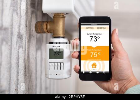 Close-up Of Person's Hand Adjusting Temperature Of Thermostat Using Cellphone At Home Stock Photo