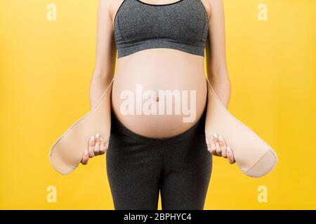 Close up of pregnant woman putting on a bandage at yellow background. Copy space. Orthopedic abdominal support belt concept. Stock Photo