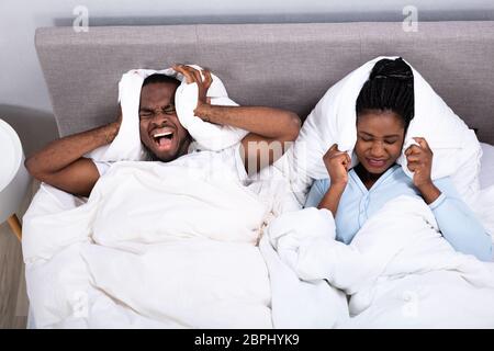 Young African Couple Disturbed By Noise Covering Their Ears With Pillow On Bed Stock Photo