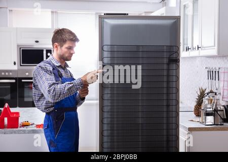 Serviceman In Overall Working On Fridge With Wrench In Kitchen Stock Photo