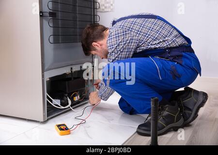 Side View Of A Male Repairman Checking Refrigerator With Digital Multimeter At Home Stock Photo