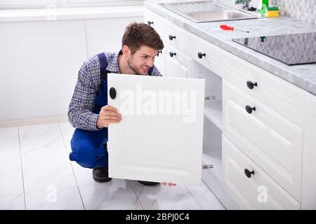 Smiling Handyman Fitting A New Door Under The Kitchen Worktop Stock Photo