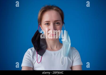 Portrait of funny woman with two medical face mask hanging on ears, looking at camera in room with blue wall at home. Self isolation, prevention Stock Photo