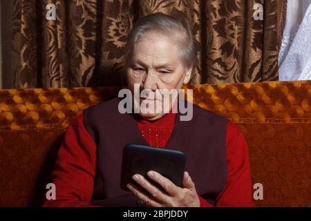 Old woman reading ebook on digital tablet device sitting in her sofa Stock Photo