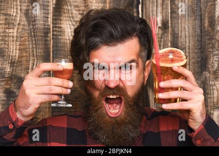 Barman with beard and angry face holds two glasses with alcohol, non alcoholic cocktail. Man and cocktails on wooden background. omplexity of choice concept. Hipster can not choose short or cocktail. Stock Photo