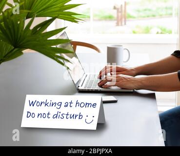 Folded paper Card standing on a table saying Working at Home - Do Not disturb. Hands of a person working on a laptop computer with coffee mug and a mo Stock Photo
