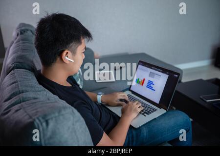 Asian businessman using wireless earphones and using computer notebook working from home.He is prepare graphs and information for online meetings at h Stock Photo