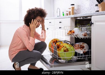 Worried Young Woman Looking At The Dirty Colorful Plates Arranged In The Dishwasher Stock Photo