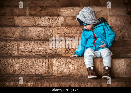 Sweet little baby boy dressed in warm clothes sitting on the steps and waiting for a parents to go in the yard for a walk, happy carefree childhood Stock Photo