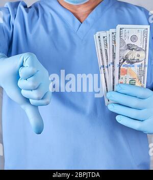 doctor in blue uniform and latex gloves keeps one hand a lot of money, the other hand shows a bad gesture, the concept of a bribe Stock Photo