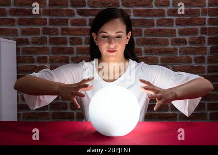 Young Woman Predicting Future With Crystal Ball On Red Desk Against Brick Wall Stock Photo