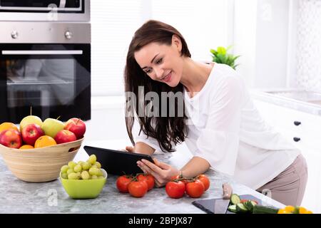 Happy Young Woman Using Digital Tablet Near Fresh Vegetables On Kitchen Counter Stock Photo