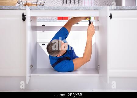 Side View Of Male Handyman Fixing Cabinet Using Screwdriver In Kitchen Stock Photo