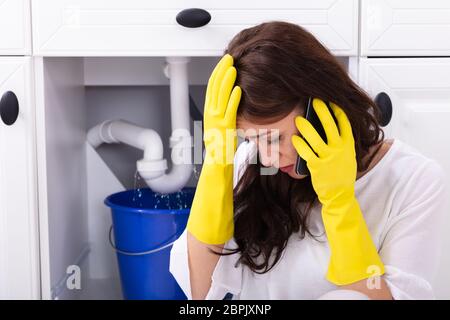 Sad Young Woman Calling Plumber In Front Of Water Leaking From Sink Pipe Stock Photo