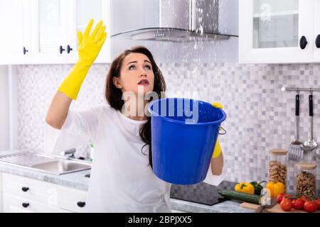 Unhappy Young Woman Collecting Water Leaking From Ceiling In Blue Bucket Stock Photo