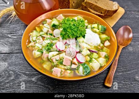 Cold soup okroshka from sausage, potatoes, eggs, radish, cucumber, greens and kvass in a bowl, bread and jug with drink on the background of wooden bo Stock Photo