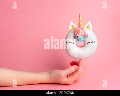 Unicorn donut on forefinger over pink background. Female hand hold trendy donut unicorn with white glaze on index finger. Copy space for text. Stock Photo