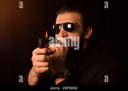 The elegant gentleman with sunglasses is burning his cigar with zippo on black background. Stock Photo