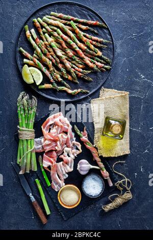 Young asparagus wrapped with bacon and roasted in the oven with garlic and sea salt served on a black plate with olive oil on a dark concrete backgrou Stock Photo