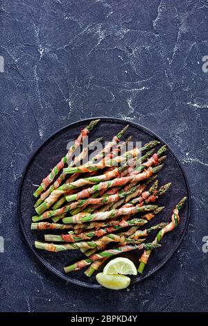Spring vegetable: asparagus wrapped in bacon and grilled with garlic and sea salt served on a black plate with olive oil, lemon on a dark concrete bac Stock Photo