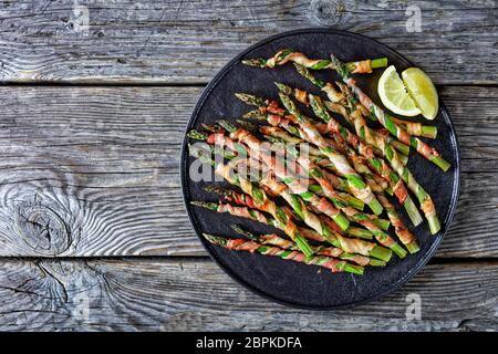 Bacon wrapped asparagus easy side dish grilled with garlic and sea salt served on a black plate lemon on an old wooden background, top view, flat lay, Stock Photo