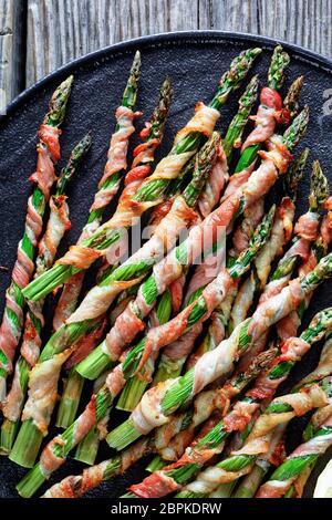 Low carb dish bacon wrapped asparagus easy side dish grilled with garlic and sea salt served on a black plate with lemon on an old wooden background, Stock Photo