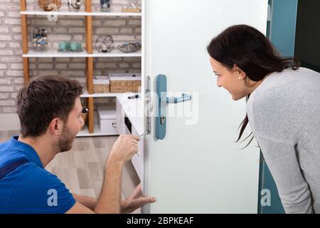 Smiling Woman Looking At Technician Fixing The Door Lock With Screwdriver At Home Stock Photo