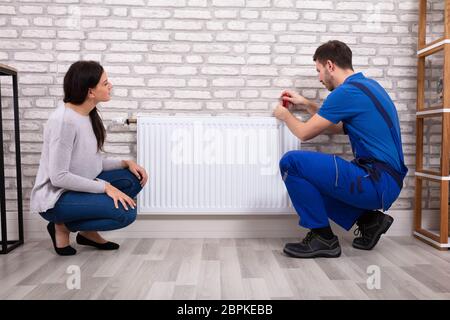 Beautiful Woman Looking At Young Male Plumber In Uniform Installing Radiator With Screwdriver Stock Photo