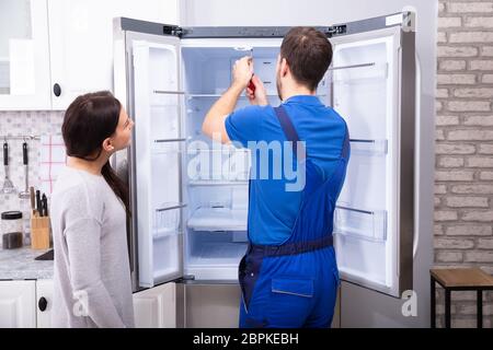 Woman Looking At Male Repairman Fixing Refrigerator With Screwdriver Stock Photo