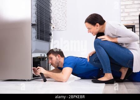 Happy Woman Looking At Technician Examining Refrigerator With Flashlight In Kitchen Stock Photo