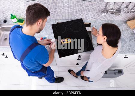 Male Repairman Installing Induction Cooker With Woman Standing In Kitchen Stock Photo