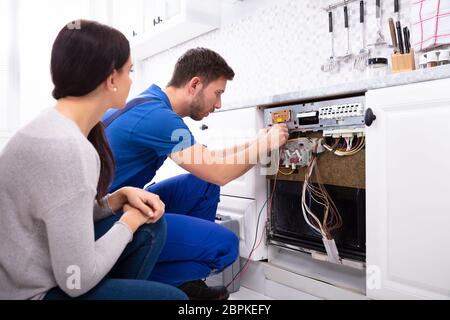 Woman Looking At Male Technician Checking Dishwasher With Digital Multimeter In Kitchen Stock Photo