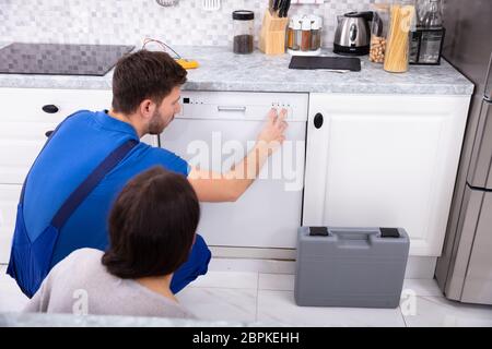 Serviceman Crouching On Kitchen Floor Pressing Button Of Dishwasher While Woman Looking At Him Stock Photo