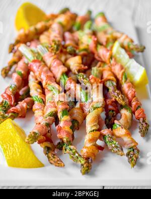 Delicious bacon wrapped asparagus fried with garlic and sea salt served on a white plate with lemon on a white wooden background, close-up Stock Photo
