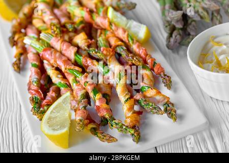 Garlic butter wrapped asparagus with bacon roasted with garlic and sea salt served on a white plate with lemon and sour cream sauce on a white wooden Stock Photo
