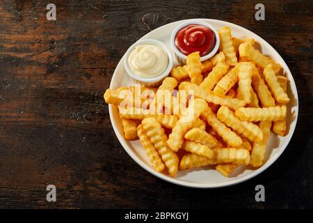 White ceramic plate of rippled french fries served with ketchup and mayonnaise, viewed from above on dark wooden background with copy space Stock Photo