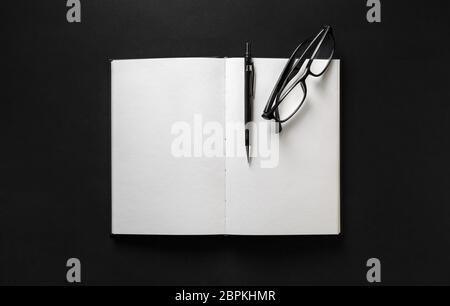 Opened blank book, glasses and pen on black paper background. Flat lay. Stock Photo