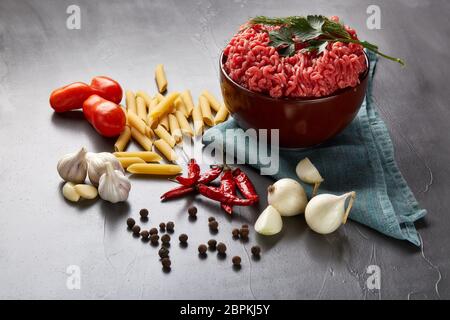 Still life with fresh forcemeat from beef in a bowl, vegetables, pasta and spices on a dark background. Stock Photo
