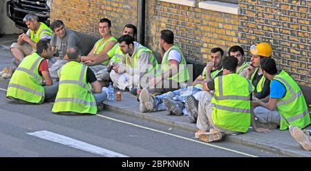 London street scene group of construction site workers sitting on public pavement outside building site wearing high vis viz jackets at lunch time UK Stock Photo