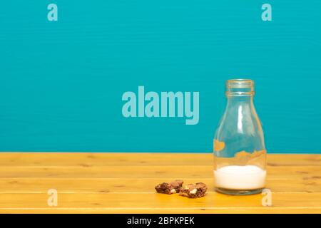 One-third pint glass milk bottle half full with fresh creamy milk and chocolate chip cookie crumbs, on a wooden table against a teal background Stock Photo