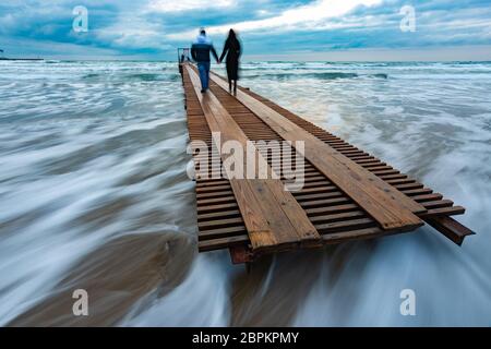 Two people go down the wooden sea pier into the distance, evening, long exposure Stock Photo