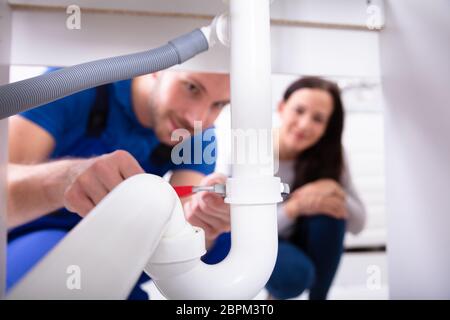 Young Woman Looking At Male Plumber Fixing Sink Pipe In Kitchen Stock Photo