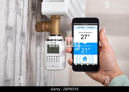 Top View Of Person's Hand Using Cellphone To Adjust Temperature Of Thermostat Stock Photo